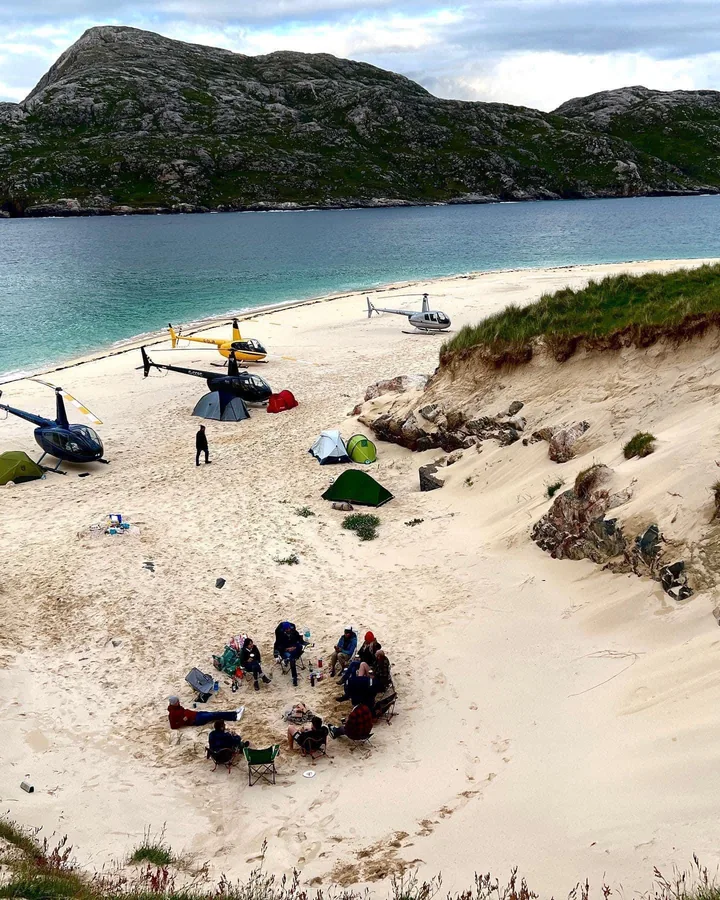 Helicopters and tents on a sandy beach beside turquoise water.